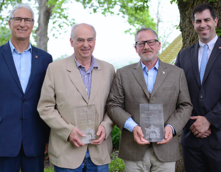 Four individuals in suits holding awards in front of a tree.