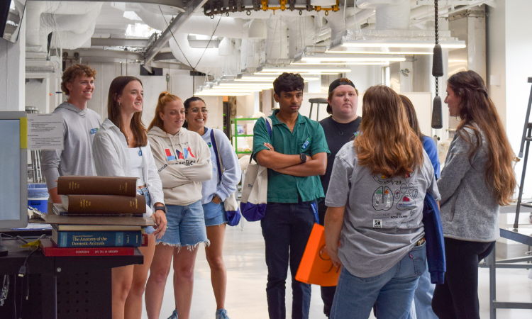 Seven new students look around their new anatomy lab, a white, industrial-looking room with metal tables and stools. Two student tour guides face away from the camera.