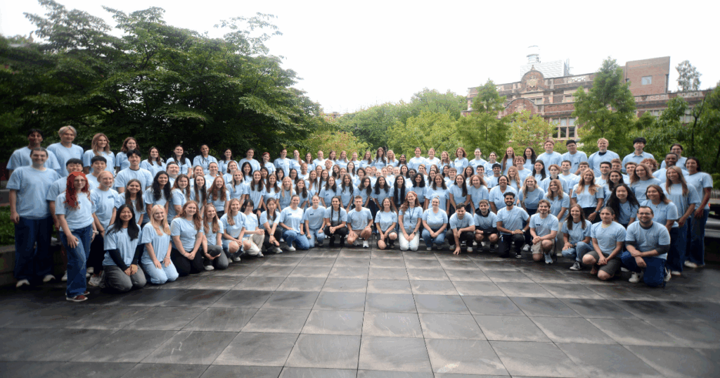 The class of 2029 wears blue orientation t-shirts and poses for a group photo. They are outside and there are trees an an academic building behind them.