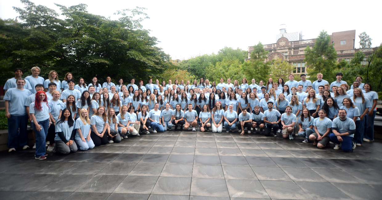 The class of 2029 wears blue orientation t-shirts and poses for a group photo. They are outside and there are trees an an academic building behind them.